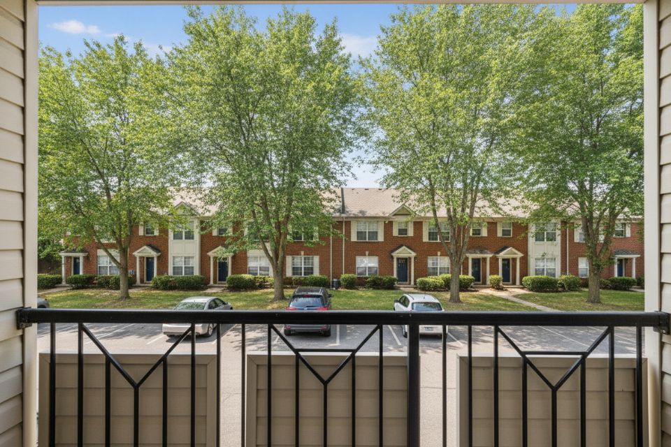 The view from a covered condominium balcony over a parking lot to trees and brick townhouses.